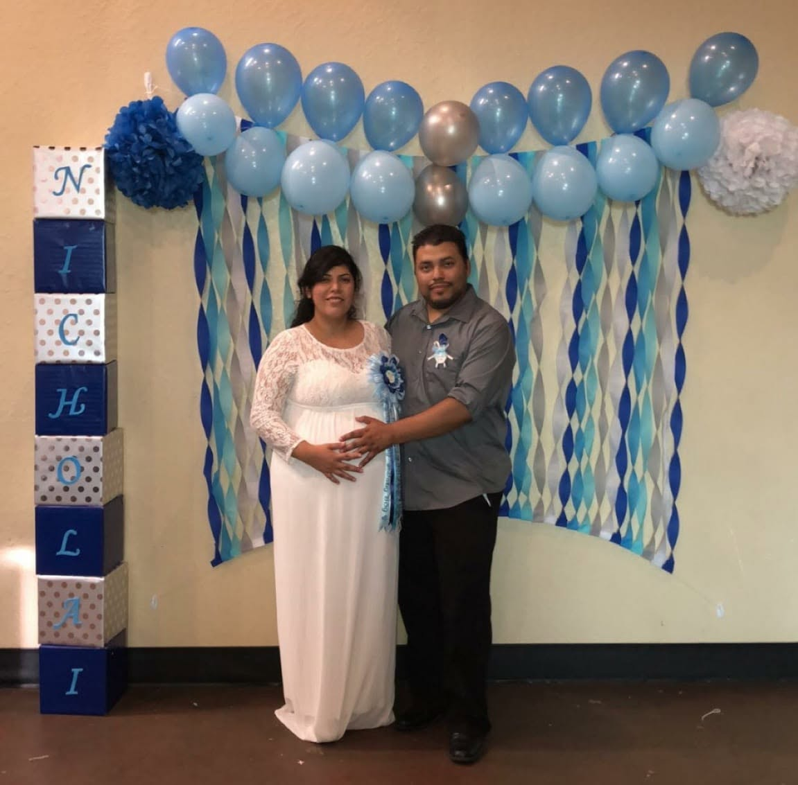 Linear garland of uniform blue and silver balloons over streamers and paper poms at a baby shower.