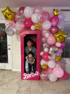 Mixed-size pink and white balloon garland with gold star foils around a doll-box photo prop at a birthday party.