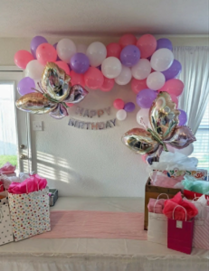 Pink, white, and purple arched garland with silver butterfly foils and birthday letters over a gift table.