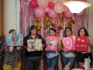 Guests hold paintings before a pink balloon garland and fringe backdrop at a friends' seasonal celebration.