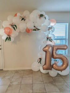 White and blush organic balloon garland with florals beside rose-gold number fifteen on a balloon base.