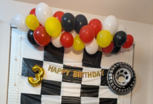 Uniform red, yellow, black, and white balloon garland above a checkered-flag backdrop for a third birthday.