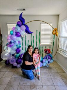 Gold hoop partially covered in purple, lavender, and teal balloons with a mermaid tail topper, indoors.