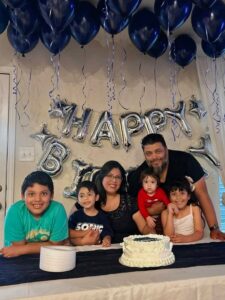 Family with cakes under navy ceiling balloons and a silver happy birthday letter banner.