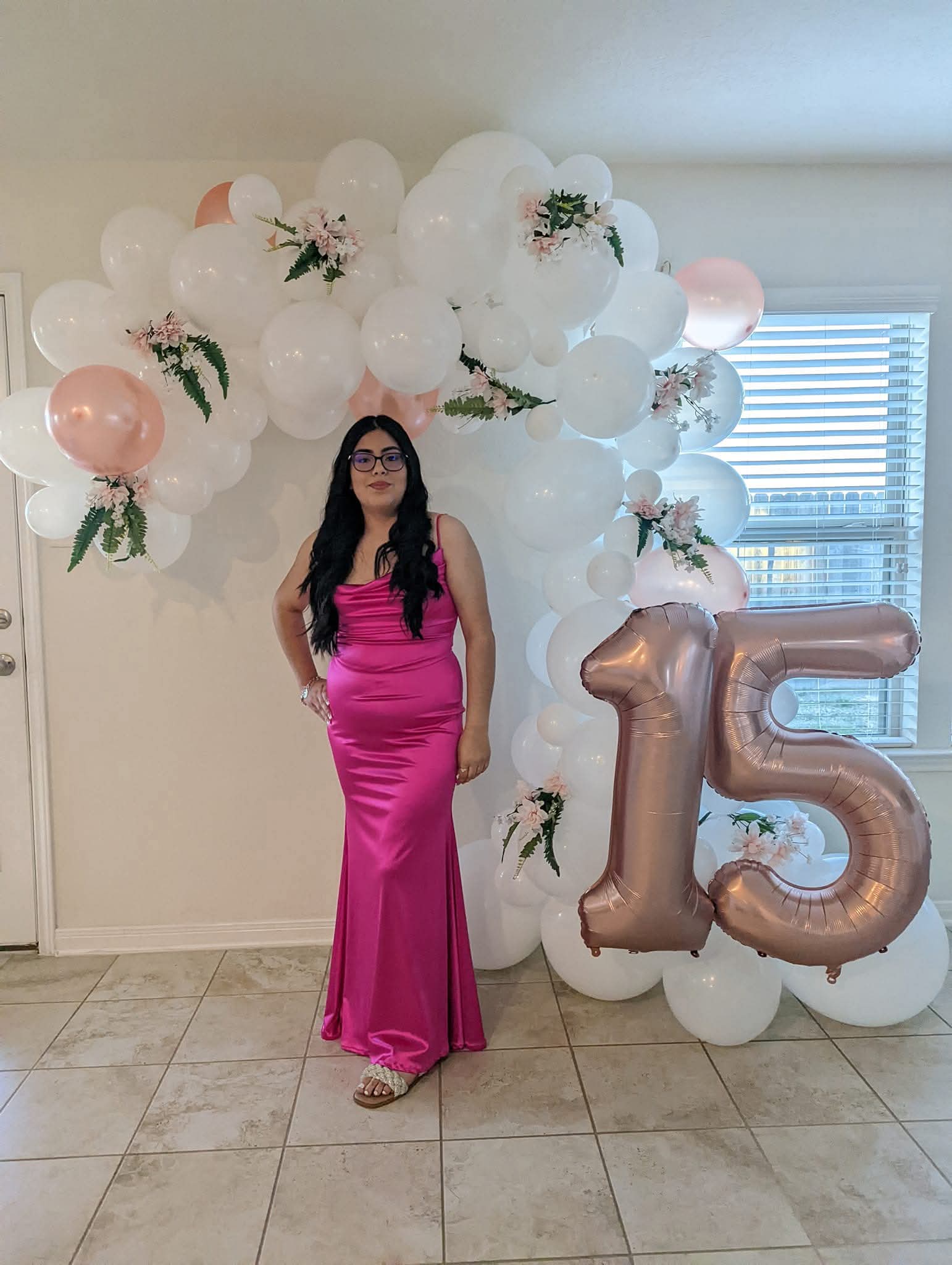 Young woman in a pink gown beside a white floral balloon garland with rose gold number balloons.