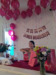 Pink helium balloons with ribbons on the ceiling above a happy birthday banner and fresh flowers.