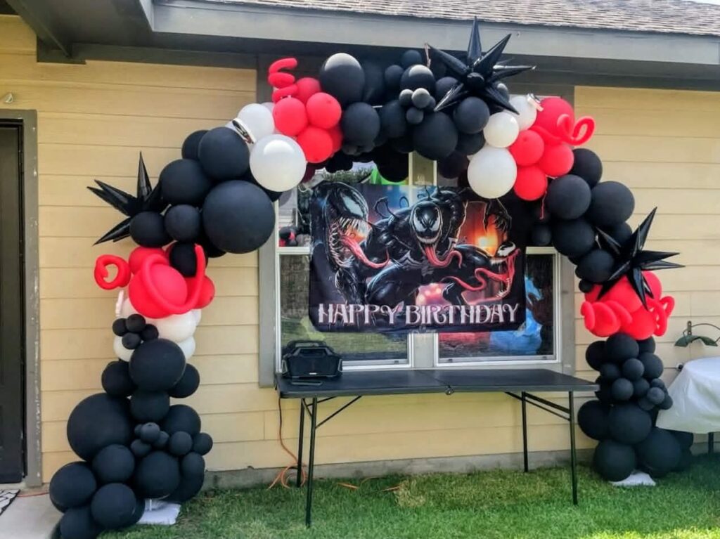 Black, red, and white organic balloon arch with star foils framing an outdoor birthday backdrop table.