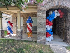 Red, white, and blue balloon columns and a doorway garland decorating a brick front porch.