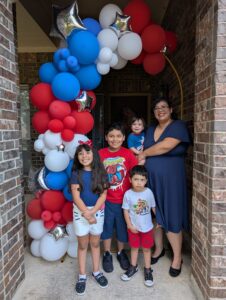 Family posing under a red, white, and blue balloon garland framing a front doorway.