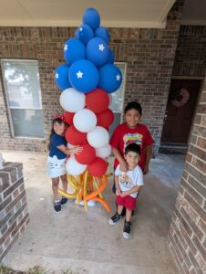 Children standing beside a stacked flag-styled balloon column on a front porch.