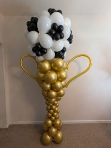 Gold balloon column sculpted as a trophy with handles and a soccer-ball patterned balloon top.