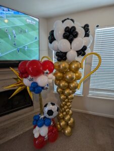 Gold trophy balloon sculpture with black and white top beside a patriotic soccer accent piece.