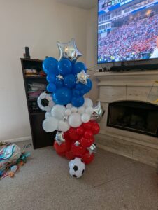 Red, white, and blue balloon column with soccer ball balloons and silver star foils indoors.