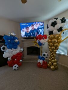 Three balloon columns in red, white, blue, gold, and black with soccer ball balloons in a living room.