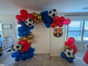 Organic balloon arch in royal blue, red, and gold with soccer ball balloons framing an interior doorway.