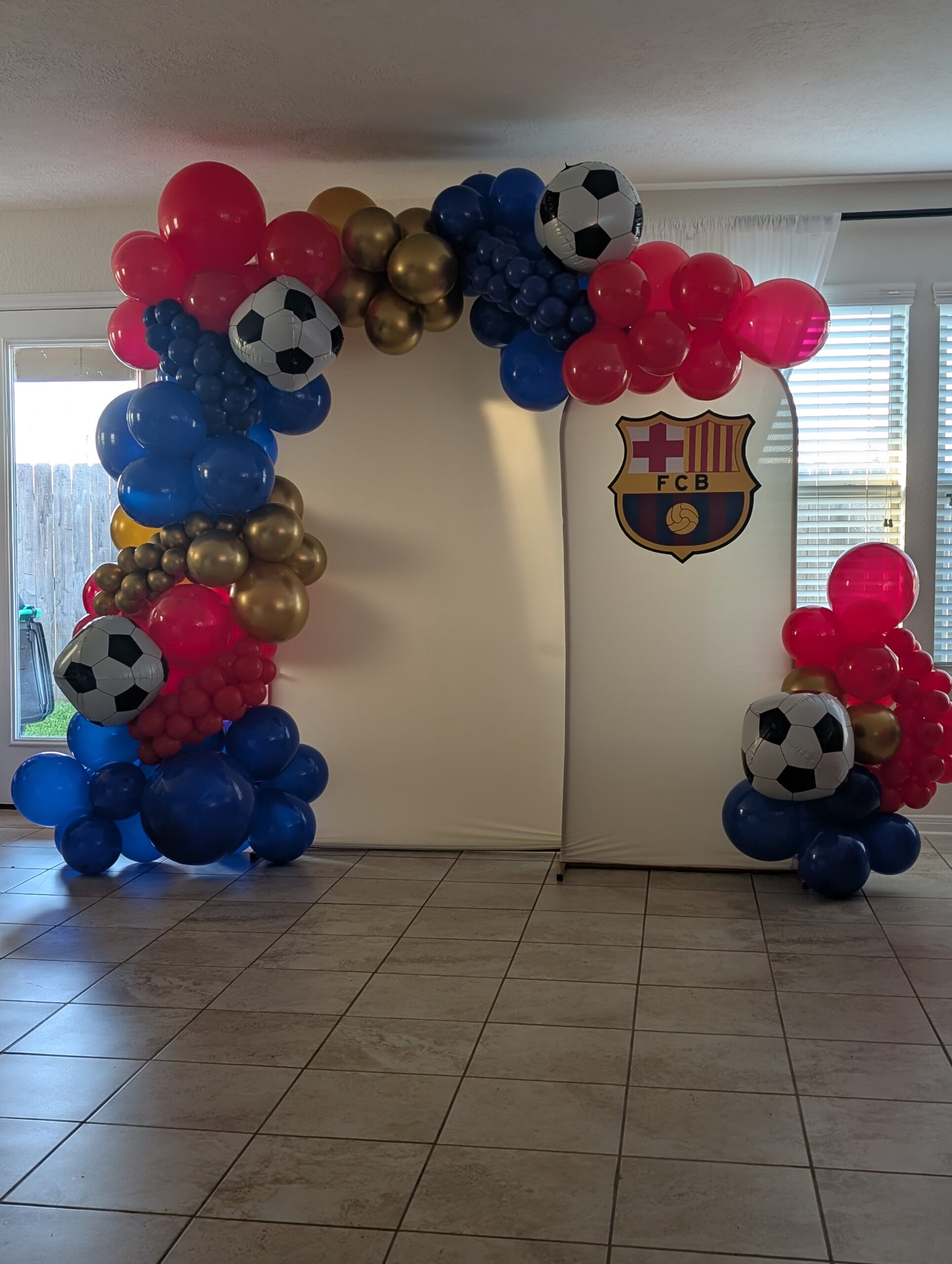 Round top backdrop panel dressed with pink, blue, and gold organic balloon garland and soccer ball balloons.