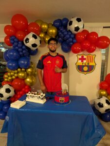 Guest of honor beside a blue, red, and gold soccer balloon garland at a birthday cake table.