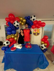 Two guests in front of a soccer themed balloon corner garland at a birthday cake table.