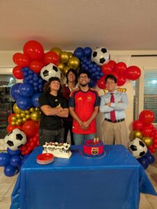 Four guests posing with birthday cakes under a red, blue, and gold soccer balloon garland.